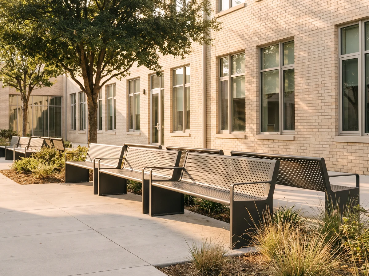 A row of charcoal steel benches with perforated seats under shade trees in a school courtyard